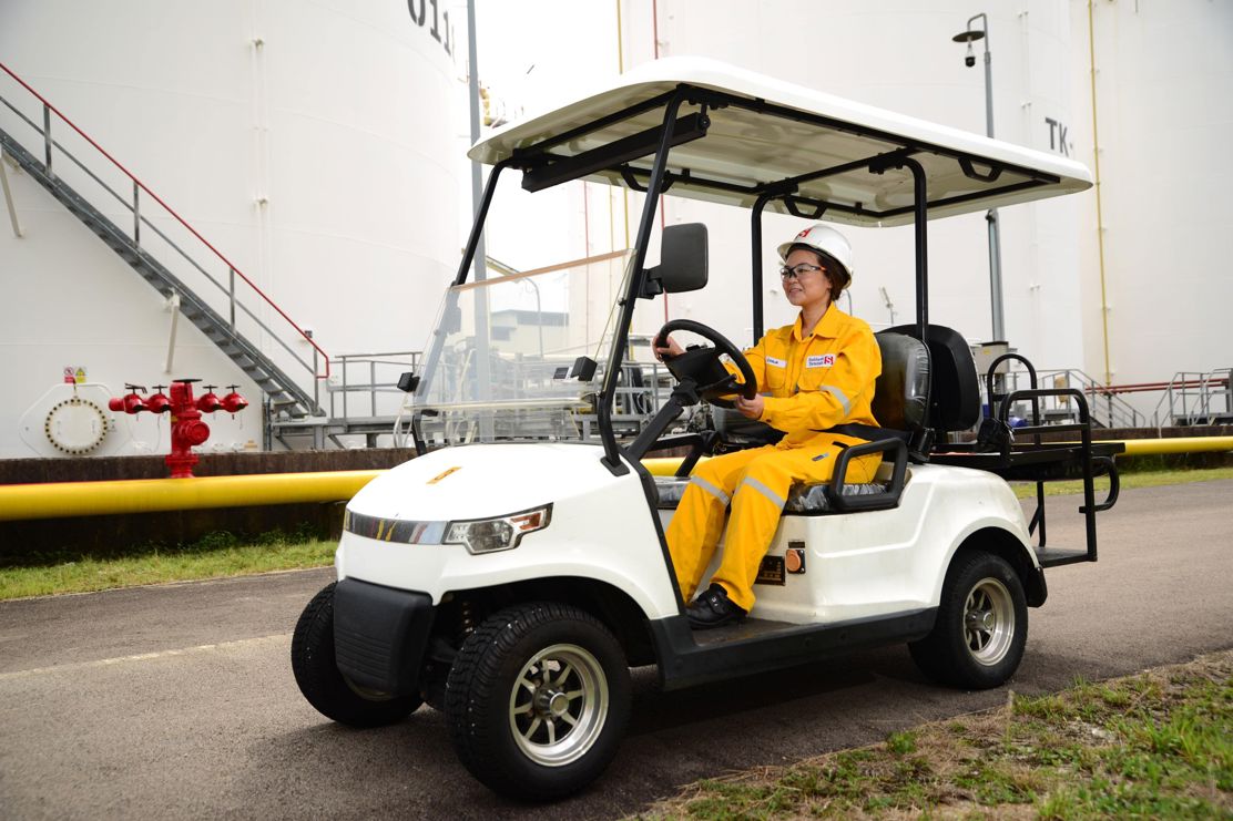 Woman in overalls and hard hat on golf buggy in front of storage tanks